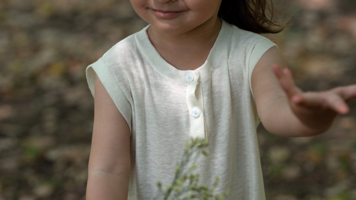 A child wearing a sleeveless cream-colored linen henley shirt, standing outdoors with a blurred forest background.
